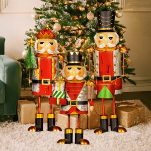 May include: Three metal nutcracker figurines standing in front of a Christmas tree. The nutcrackers are dressed in red and green uniforms with gold accents. The nutcrackers are holding various items, including a drum, a sword, and a candy cane.
