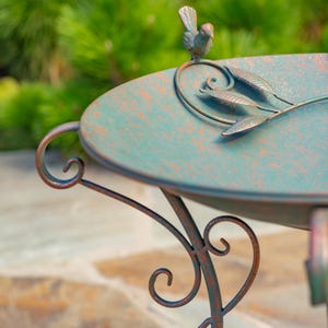 Puede incluir: Un bebedero decorativo de metal envejecido, con una cuenca verde-turquesa y soportes ornamentados con volutas. Una peque&ntilde;a escultura de p&aacute;jaro se posa sobre el borde, y detalles de hojas adornan la cuenca. El bebedero tiene un aspecto r&uacute;stico y antiguo.