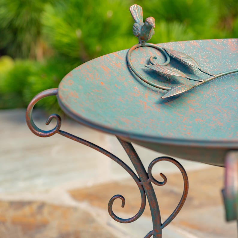 Puede incluir: Un bebedero decorativo de metal envejecido, con una cuenca verde-turquesa y soportes ornamentados con volutas. Una peque&ntilde;a escultura de p&aacute;jaro se posa sobre el borde, y detalles de hojas adornan la cuenca. El bebedero tiene un aspecto r&uacute;stico y antiguo.