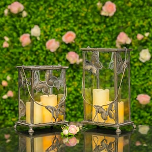 May include: Two decorative metal lanterns with butterfly designs and glass panels. Each lantern holds three pillar candles, creating a warm glow. The lanterns are set against a backdrop of green foliage and pink flowers.