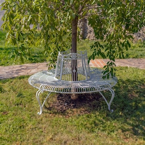 May include: A white circular metal tree bench featuring a decorative wire lattice seat and a matching vertical backrest that encircles the trunk of a tree. The bench is supported by curved metal legs and is positioned on a patch of green grass.