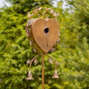 May include: A heart-shaped birdhouse with a copper finish and a small opening for birds to enter. The birdhouse is attached to a metal stand with decorative leaves and bells.