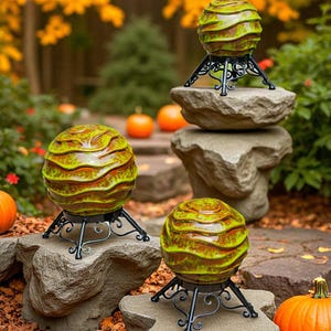 May include: Three spherical ceramic objects with a glossy, wavy green and brown glaze. Each sphere rests on a black metal stand featuring intricate, curved scrollwork legs. The objects are arranged at different heights on stone surfaces.