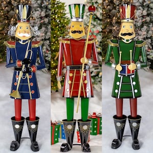 May include: Three metal Christmas nutcracker soldier figurines in blue, red, and green. Each soldier has a tall hat, a mustache, and holds a musical instrument or staff. The soldiers stand on black boots.