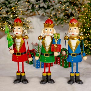 May include: Three festive metal nutcracker figurines, each with a different colored uniform: red, green, and blue. They are holding a small Christmas tree, a gift box, and a wreath, respectively.