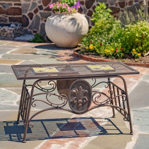 May include: A rectangular bronze-colored metal table featuring a slatted top with two inset square decorative tiles. The table has curved metal legs and a central oval medallion with a raised fleur-de-lis design on the front support frame.