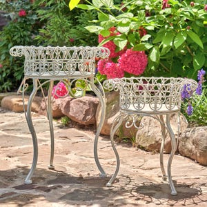 May include: Two white metal plant stands with ornate designs. The stands have a round top and three legs. The stands are in a garden setting.