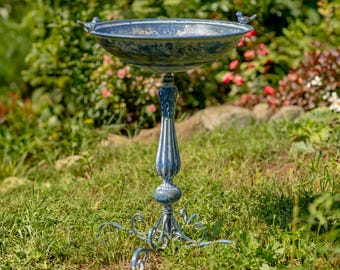 May include: A blue metal bird bath with a decorative pedestal base. Two small bird figurines are perched on the rim. The bird bath is set in a garden with green grass and foliage.