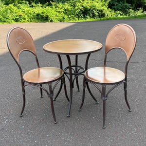 May include: A bistro-style outdoor dining set with a round table and two chairs. The table has a light brown wooden top and a dark metal base. The chairs have brown leather seats and backs, with a decorative emblem on the backrests. The set is designed for outdoor use.