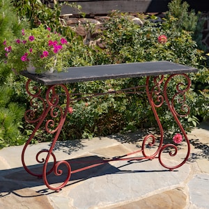 May include: A rectangular console table featuring a dark, weathered metal top and a red, scrollwork metal base with ornate, swirling legs. A white pot filled with vibrant pink flowers sits on the left side of the table surface.