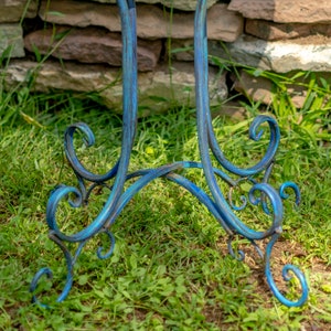 May include: A close-up of the base of a blue metal decorative stand. The stand has a scrolled design and is painted in shades of blue. The stand is on a bed of green grass.