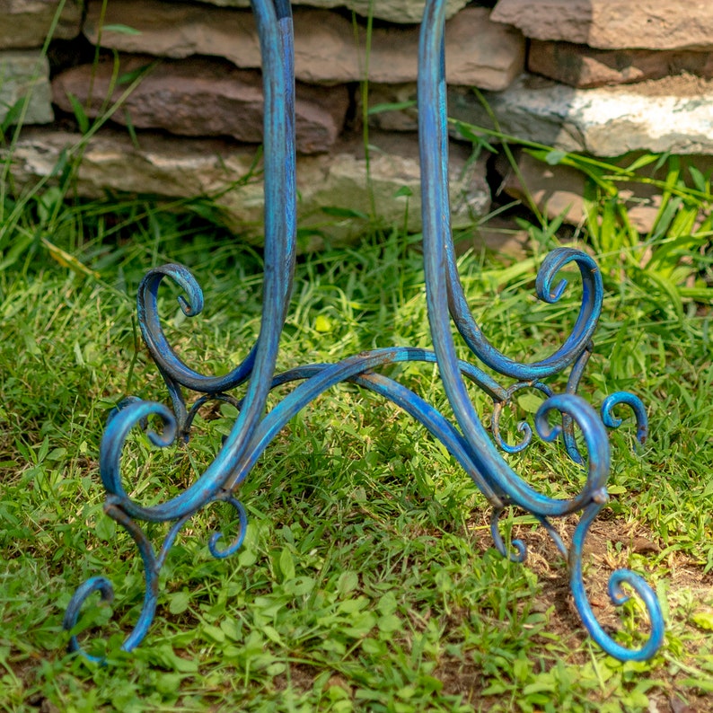 May include: A close-up of the base of a blue metal decorative stand. The stand has a scrolled design and is painted in shades of blue. The stand is on a bed of green grass.