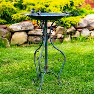 May include: A decorative, antique-style bird bath with a teal finish. The metal stand features ornate scrollwork and supports a shallow basin. Three small bird figurines perch on the rim. The bird bath is set on a green lawn with a stone wall backdrop.