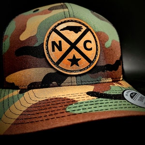 May include: A camouflage trucker hat with a brown leather patch featuring the outline of North Carolina and the letters "NC" with a star in the center.