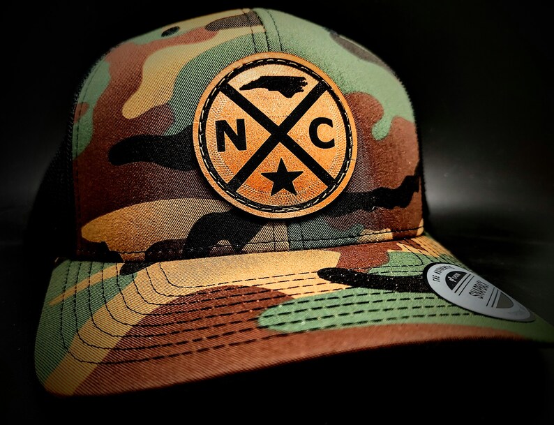 May include: A camouflage trucker hat with a brown leather patch featuring the outline of North Carolina and the letters "NC" with a star in the center.