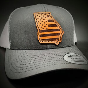 May include: A gray and white trucker hat with a brown leather patch shaped like the state of Georgia. The patch features a red, white, and blue American flag design.