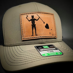 May include: A green and black trucker hat with a brown leather patch featuring a black silhouette of a pirate holding a spear and a heart. The patch has the text "112RE" and "RECYCLED FABRICS" on it.
