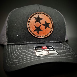 May include: A black and gray trucker hat with a brown leather patch featuring three stars. The patch is stitched around the edges. The hat has a black and gray mesh back and a black adjustable strap with a black plastic buckle. The strap has a white label with the text "EST. 1970 RICHARDSON ORIGINAL TRUCKER 112 ADJUSTABLE".