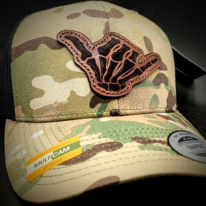 May include: A camouflage trucker hat with a black mesh back. The hat has a brown leather patch with a skeleton hand making a shaka sign. The hat has a label that says "MULTICAM".