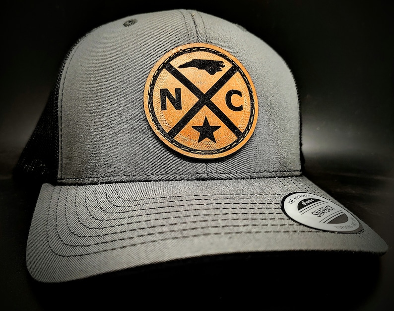 May include: A gray and black trucker hat with a brown leather patch featuring a silhouette of North Carolina and the letters "NC" in a circle.