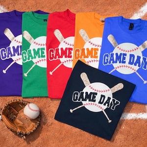 May include: A collection of baseball-themed t-shirts in various colors, including purple, green, red, orange, blue, and navy. Each shirt features a baseball graphic with the words "GAME DAY". A baseball glove and ball are also present.