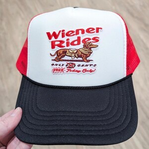 以下が含まれることがあります： 黒いつばのある白と赤のトラッカーハット。ハットの前方には、「Wiener Rides」と刺繍された赤と白のパッチがあり、サドルをつけたダックスフントが描かれています。パッチには、「Only 20 Cents Free Today Only!」の文字も書かれています。