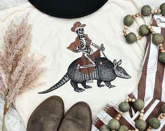 May include: A white t-shirt with a graphic of a skeleton wearing a cowboy hat riding an armadillo. The armadillo is brown and black with a saddle on its back. The skeleton is holding the reins in its hands.