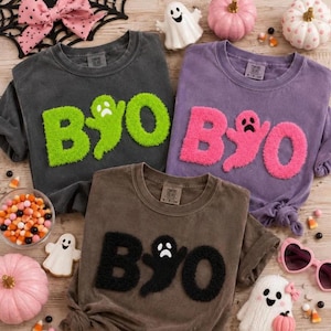 May include: Three crewneck t-shirts in charcoal, purple, and brown, each featuring fuzzy chenille lettering that spells BOO with a ghost replacing the letter O. The charcoal shirt has lime green letters, the purple has pink, and the brown has black.