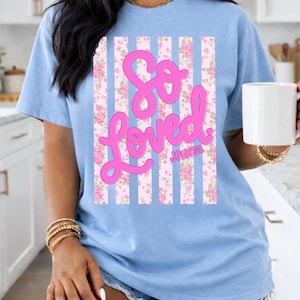 May include: Pale blue t-shirt with the words "So Loved" in pink script. The design features a floral pattern with pink roses and vertical white stripes. The text "JOHNSI6" is below the word "Loved".