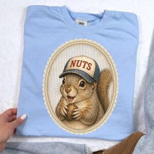 May include: Light blue sweatshirt featuring a detailed illustration of a squirrel wearing a baseball cap with the word "NUTS" on it. The squirrel is holding a nut. The graphic is inside an oval frame.