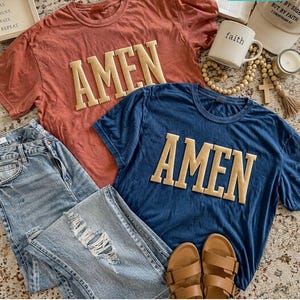 May include: Two short-sleeved t-shirts, one coral and one navy blue, each with the word "AMEN" in a tan, collegiate-style font. The shirts are paired with light-wash denim jeans, brown sandals, and religious accessories.