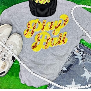 May include: A gray t-shirt with a yellow and red sequin softball patch that says "Play Ball". The shirt is laying on a green surface with a white beaded necklace, a black baseball cap with a yellow and red sequin softball patch, and a pair of white sneakers.