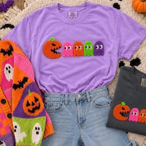 May include: A lavender short-sleeve t-shirt featuring a fuzzy orange pumpkin character and four colorful ghost characters in pink, orange, green, and purple. The design is centered on the chest. A dark gray t-shirt with an identical design is also visible.