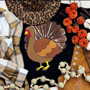 Puede incluir: Una camiseta negra con un gran aplique de pavo con lentejuelas. La imagen también presenta un sombrero con estampado de leopardo, decoraciones de calabazas naranjas, una camisa a cuadros y una prenda de pana marrón, lo que sugiere un tema de otoño o Acción de Gracias.