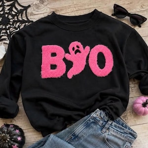 May include: A black long-sleeve crewneck sweatshirt featuring the word BOO in fuzzy, bright pink chenille letters, with the letter O replaced by a ghost shape. The sweatshirt is displayed alongside blue denim jeans, sunglasses, and pumpkins.