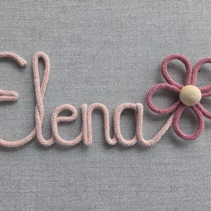 Könnte beinhalten: Ein personalisiertes Namensschild mit dem Namen Elena aus hellrosa gestrickter Kordel, ergänzt durch eine dunkelrosa gestrickte Blume mit einer hellen Holzperle in der Mitte. Die Schreibschrift ist mit der Blume zu einer Wanddeko verbunden.