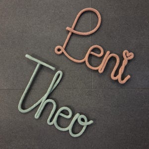 Könnte beinhalten: Zwei Namen, "Theo" und "Leni", in Garn geschrieben. Der Name Theo ist in grünem Garn und der Name Leni ist in rosafarbenem Garn.