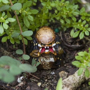 May include: A small, brown and black Predator action figure with red eyes and a black mouth is hidden in a bed of green plants. The figure is standing on a bed of brown dirt.