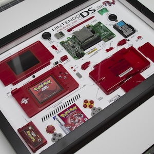 Op de afbeelding: Een gedemonteerde Nintendo DS Lite in een rood-wit kleurenschema, tentoongesteld in een zwart frame. De afzonderlijke componenten zijn gelabeld, inclusief het LCD-scherm, de aan/uit-knop en het moederbord. De gamecartridge-sleuf is zichtbaar, samen met een Pokemon Ruby Version gamecartridge.