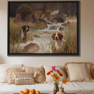 May include: A framed painting depicts two dogs in a stream. One dog is black and white spotted, the other is white and brown. Tall grass surrounds the dogs and the water. The background shows a forest and rocks. The painting is in a black frame.