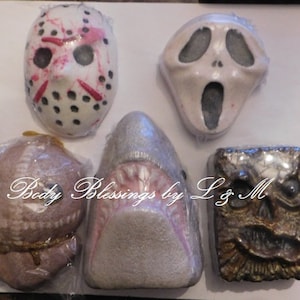 May include: An assortment of bath bombs in various shapes, including a hockey mask, a Scream mask, a shark, a doll, and a book. The bath bombs are wrapped in clear plastic. The text "Body Blessings by L & M" is visible.