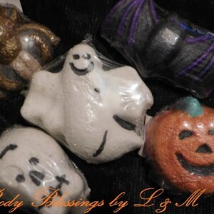 May include: A collection of five bath bombs wrapped in clear plastic, featuring a gold spider, a black bat with purple wings, a white ghost with a smiling face, a white skull, and an orange pumpkin with a carved jack-o'-lantern face.