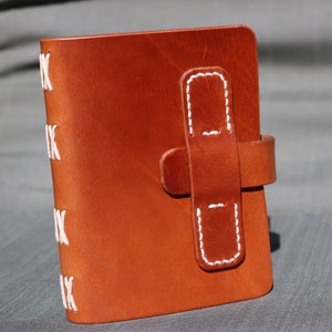 May include: A brown leather journal with white stitching around the edges and a leather strap with white stitching. The journal is closed and standing upright on a gray surface.