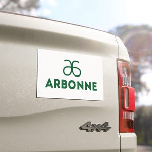 May include: A white rectangular magnet displaying the word "ARBONNE" in dark green block letters, with a green logo above. The magnet is attached to the rear of a light-coloured vehicle.