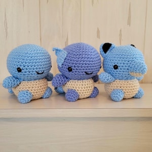 May include: Three crocheted stuffed toys in the shape of a blue and beige Pokemon character. The toys are sitting on a light wood surface.