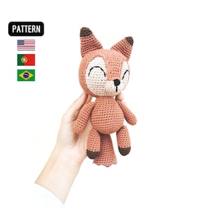 Pode incluir: Um brinquedo de pelúcia de raposa de crochê com orelhas marrons e um corpo marrom claro. A raposa tem um rosto branco com olhos pretos e um nariz preto. A raposa está sorrindo.