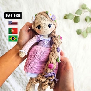 Pode incluir: Boneca de crochê com cabelo longo castanho, um vestido roxo e rosa e uma coroa de flores. A boneca está segurando uma trança de cabelo com flores.