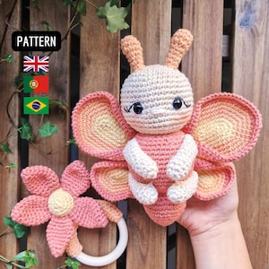 Pode incluir: Um brinquedo de borboleta de crochê com asas rosa e amarelas e um corpo branco. O brinquedo tem olhos pretos e uma pequena flor de crochê presa a um anel de madeira.