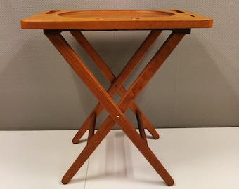 Puede incluir: Una mesa plegable de madera con una bandeja en la parte superior. La mesa tiene un acabado de madera natural y un diseño sencillo.