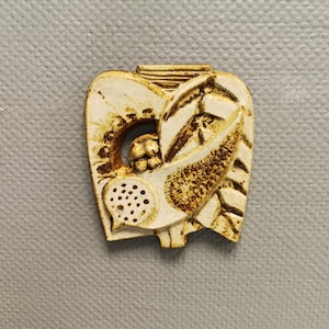 May include: A white ceramic pendant with a brown glaze depicting a stylized bird with leaves and berries. The pendant has a textured surface and a hole at the top for hanging.